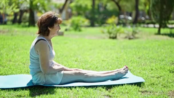 中年妇女坐在公园的瑜伽垫上伸腿 — 图库视频影像