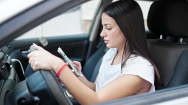 Young beautiful hispanic woman using smartphone sitting on car at street