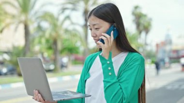 Genç Çinli bir kadın parkta dizüstü bilgisayar kullanarak akıllı telefondan konuşuyor.