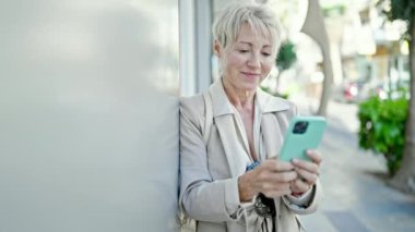 Orta yaşlı sarışın kadın sokakta akıllı telefon kullanarak gülümsüyor.