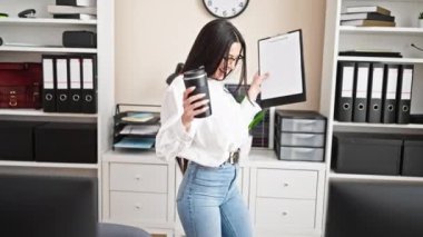 Genç, güzel İspanyol iş kadını kahve içiyor, elinde pano, ofiste dans ediyor.