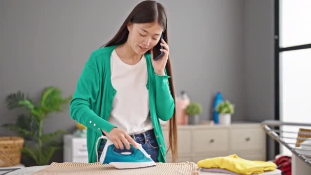 若い中国人女性が洗濯室でスマートフォンのアイロン掛け服について話す — ストック動画