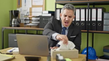 Genç İspanyol iş adamı ofiste hapşırırken dizüstü bilgisayar kullanıyor.