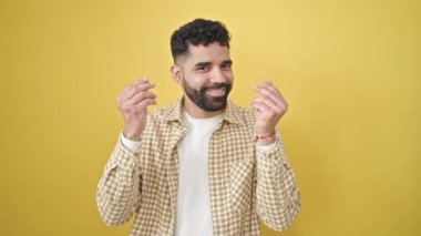 Young hispanic man smiling confident doing spend money gesture over isolated yellow background