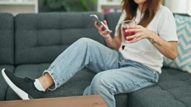 年轻美丽的他的惊慌失措的女人用智能手机躺在沙发上 在家里端着一杯茶 — 图库视频影像