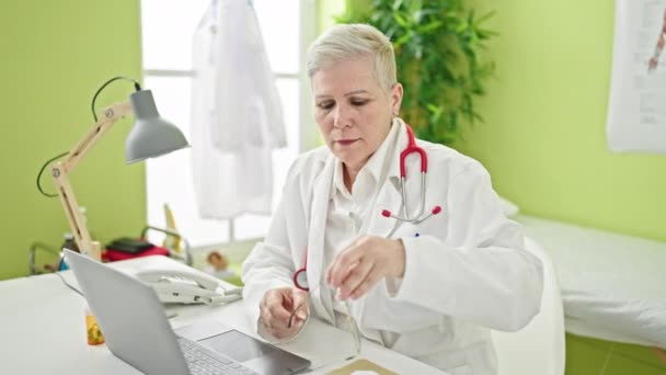 中年灰发女医生 手持笔记本电脑 戴着眼镜 在诊所微笑 — 图库视频影像