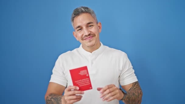 孤立した青色の背景にスイスのパスポートを保持する自信を持って微笑む若いヒスパニックの男 — ストック動画