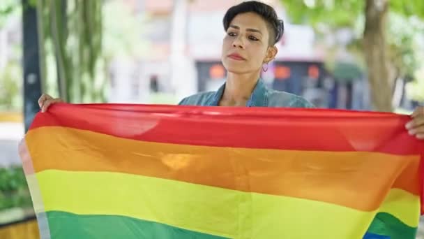 公園で虹の旗を掲げ深刻な表現で立っている若い美しいヒスパニックの女性 — ストック動画