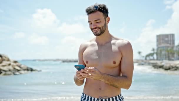 Молодий Іспаномовний Чоловік Турист Купальнику Використовуючи Смартфон Посміхаючись Пляжі — стокове відео