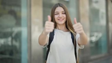 Sırt çantası takan genç ve güzel bir kız sokakta baş parmak hareketi yapıyor.