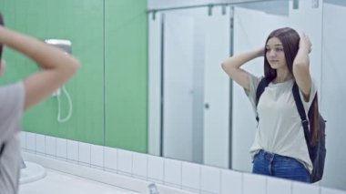 Genç güzel kız öğrenci aynaya bakıyor banyoda saçlarını tarıyor.