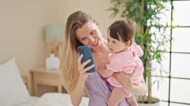 Anne ve kızı yatakta oturmuş, yatak odasında akıllı telefondan selfie çekiyorlar.