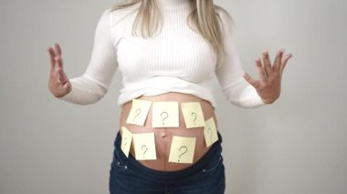 Genç hamile kadın, karnındaki soru işaretlerine işaret ederek izole edilmiş beyaz arka planda bebek cinsiyeti sorduğunu hatırlatıyor.