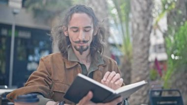 Young hispanic man reading book sitting on table at coffee shop terrace