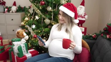 Genç hamile kadın akıllı telefon kullanıyor. Evde Noel 'i kutlamak için kahve içiyor.