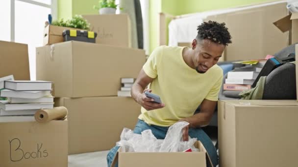African American Man Unpacking Cardboard Box Using Smartphone New Home — Stock Video