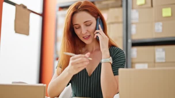 年轻的红头发女性电子商务商人在办公室里用智能手机在笔记本上写字 — 图库视频影像