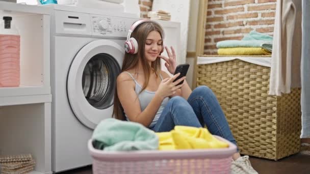 洗濯室で洗濯機を待っている音楽を聞いている若い美しい女の子 — ストック動画