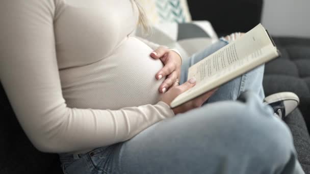 Молодая Беременная Женщина Читает Книгу Касаясь Живота Дома — стоковое видео