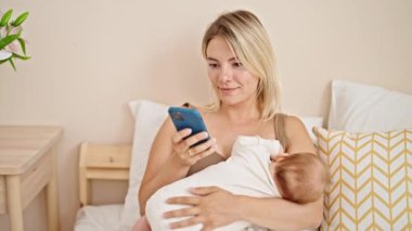 Anne ve kızı yatak odasında akıllı telefon kullanarak bebeği emziriyorlar.