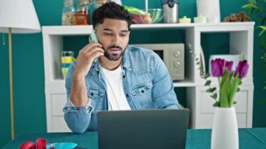 Afro-Amerikalı adam yemek odasında dizüstü bilgisayar kullanarak akıllı telefondan konuşuyor.