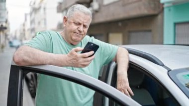 Orta yaşlı, kır saçlı, akıllı telefon kullanan bir adam caddede duruyor.
