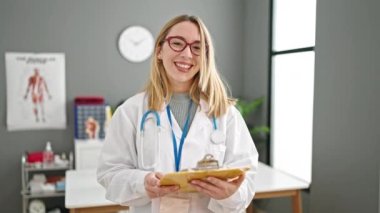 Genç sarışın doktor gülümseyerek klinikte kendine güvenen bir pano tutuyor.