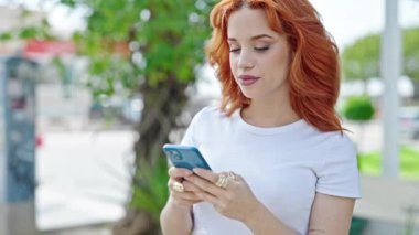 Genç kızıl saçlı kadın parkta akıllı telefon kullanıyor.