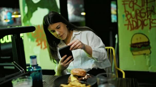 若い美しいヒスパニック系の女性がレストランでスマートフォンでおいしいハンバーガーの写真を撮る — ストック動画