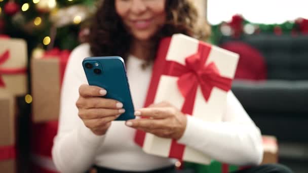 家でクリスマスツリーによって床に座っているスマートフォンを使用して中世のパニック女性 — ストック動画