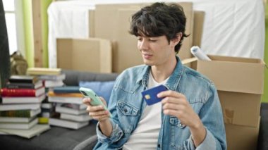 Genç İspanyol adam akıllı telefon ve kredi kartıyla alışveriş yapıyor. Yeni evindeki kanepede oturuyor.