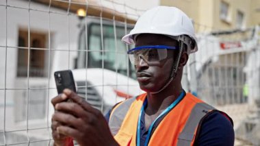 Afro-Amerikalı inşaatçı ciddi bir yüzle şantiyede akıllı telefon kullanıyor.