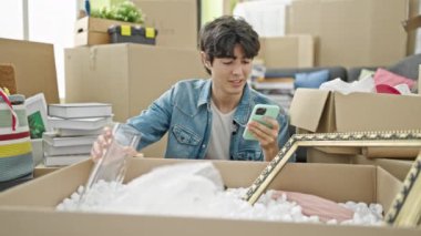 Genç İspanyol adam yeni evinde karton kutuları açmak için akıllı telefon kullanıyor.
