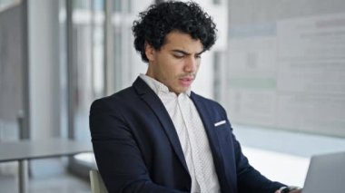 Laptop kullanan genç Latin iş adamı ofise üzgün bakıyor.