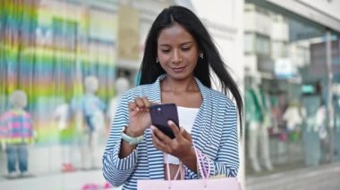 Afro-Amerikalı kadın akıllı telefon kullanıyor. Alışveriş torbalarını sokakta tutuyor.