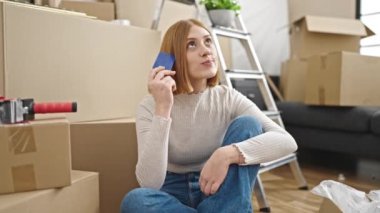 Genç sarışın kadın yeni evinde kredi kartı taşıdığını düşünerek yerde oturuyor.