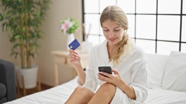 年轻的金发女人穿着浴衣 在卧室里带着信用卡和智能手机购物 — 图库视频影像