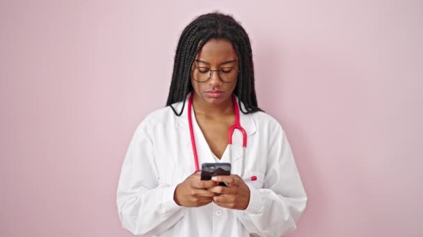 アフリカ系アメリカ人の女性医師がピンクの背景にスマートフォンを使って微笑む — ストック動画