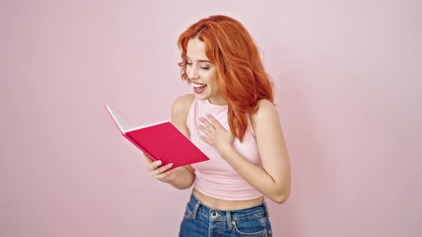 Молода Руда Жінка Читає Книгу Посміхаючись Над Ізольованим Рожевим Тлом — стокове відео