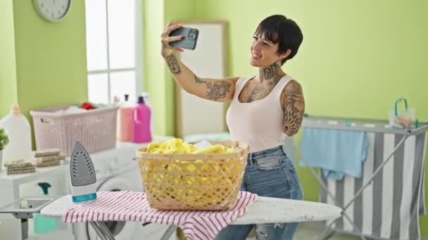 히스패닉 여성은 세탁실에서 다림질 테이블로 스마트 폰으로 셀카를 만듭니다 — 비디오
