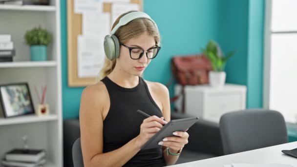 若い金髪女性ビジネスワーカーは オフィスで音楽を聴くタッチパッドに書いています — ストック動画