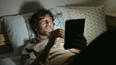 Young hispanic man using touchpad lying on bed at bedroom