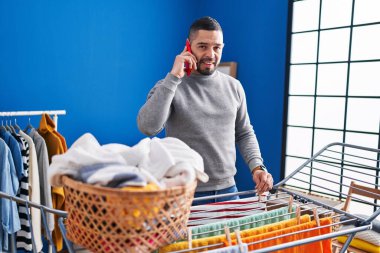 Young latin man talking on smartphone hanging clothes on clothesline at laundry room