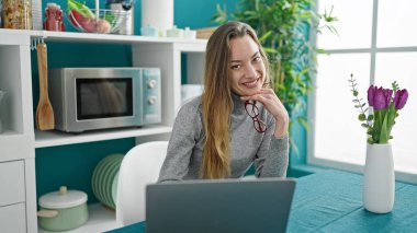 Genç beyaz kadın yemek odasında masada dizüstü bilgisayar kullanıyor.