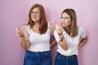 İspanyol anne ve kızı pembe arka plan üzerine gündelik beyaz tişört giyip el ve parmaklarıyla italyan jestleri yapıyorlar. 