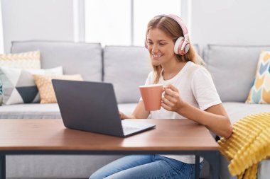 Young blonde girl watching video on laptop drinking coffee at home