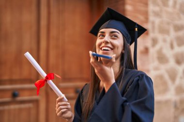 Young hispanic woman wearing graduated uniform holding diploma talking on the smartphone at university