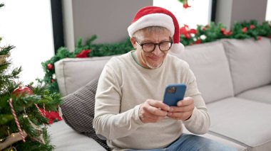 Orta yaşlı, kır saçlı adam akıllı telefon kullanıyor. Yılbaşı ağacının yanında oturuyor.