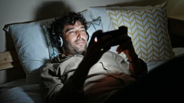Genç İspanyol adam, yatakta uzanmış akıllı telefondan video izliyor.