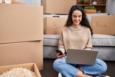 Young hispanic woman using laptop and smartphone sitting on floor at new home